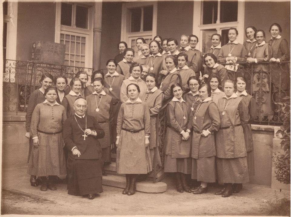 Az anyaház udvarán Engelbert lelkiigazgatóval az 1930-as években. (Margit testvér középen, a hátsó sorban áll az ajtó előtt.) Kép forrása: www.facebook.com/Szociális-Testvérek-Társasága-143677702329777